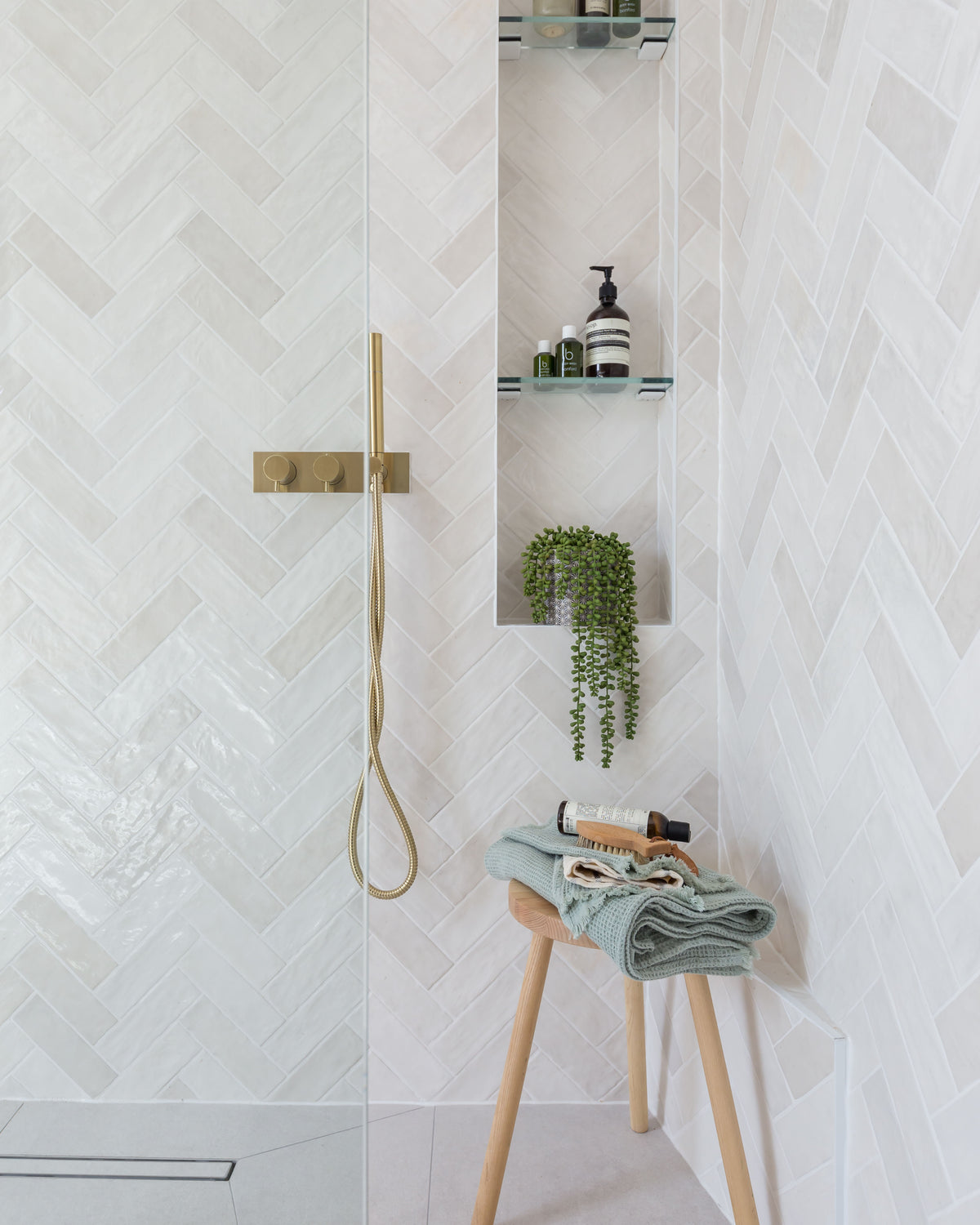 Oasis Ceramic White Gloss in a herringbone pattern across a walk-in shower with brushed brass fittings and a tiled niche