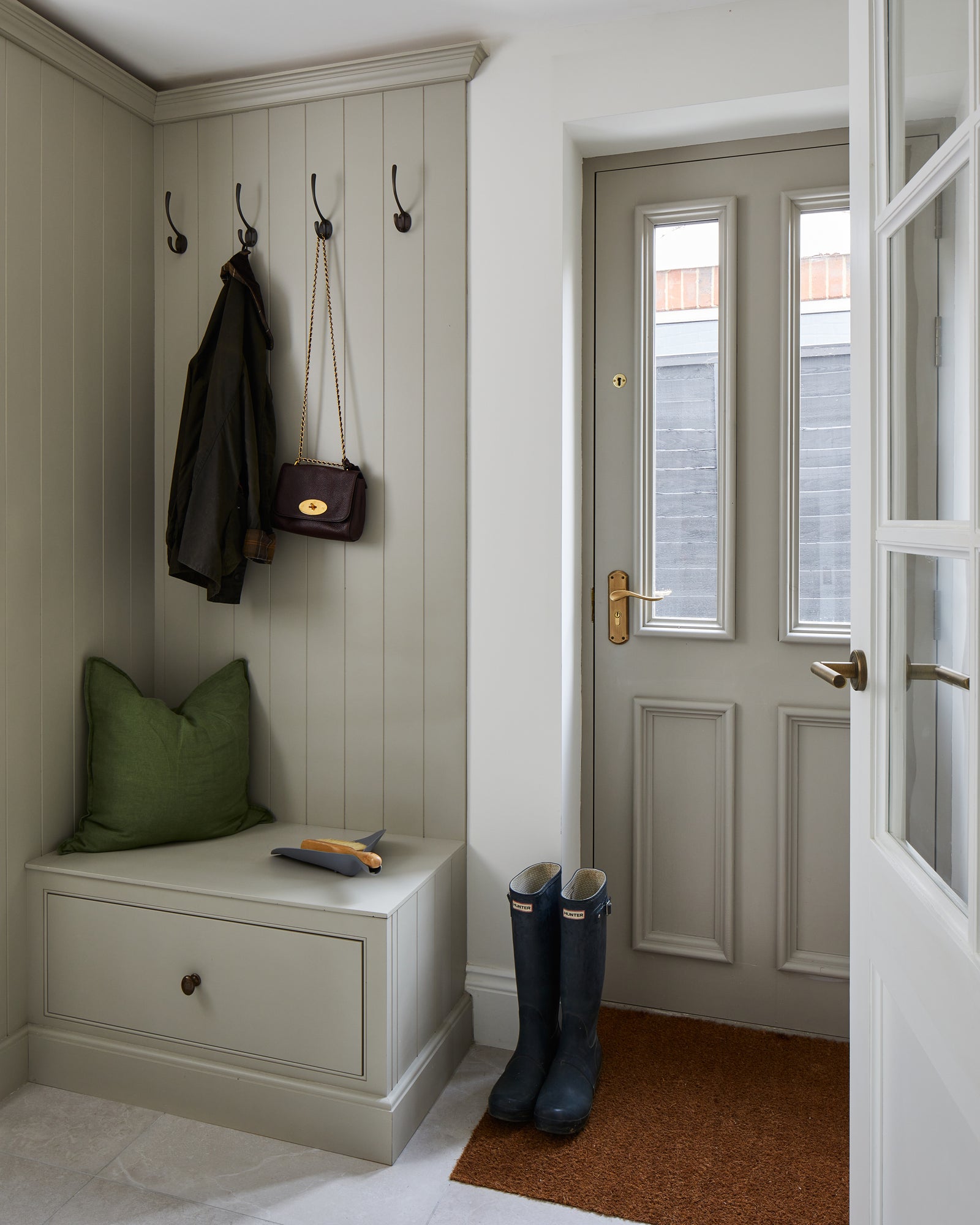 Riverside Porcelain Perla floor tiles in a panelled boot room, with a green cushioned bench, hanging waxed jacket and navy wellingtons by the back door