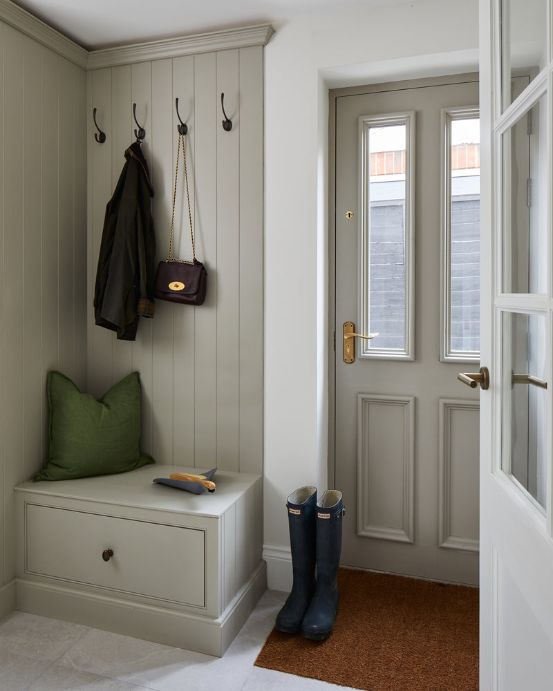 Riverside Porcelain Perla floor tiles in a panelled boot room, with a green cushioned bench, hanging waxed jacket and navy wellingtons by the back door