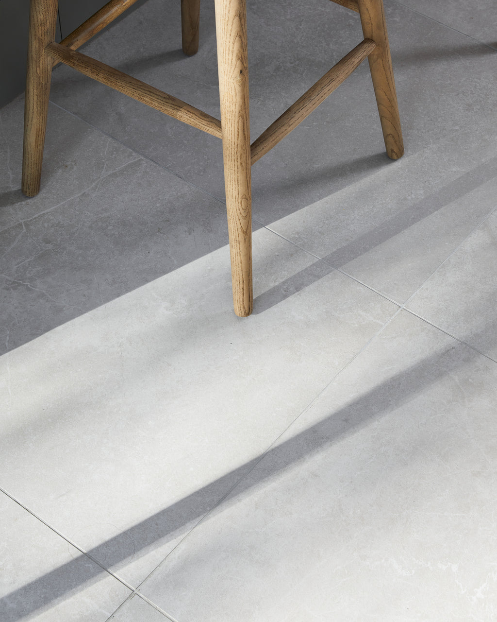 Close-up of Riverside Porcelain Perla floor tiles under an oak bar stool, raking sunlight picking out the stone texture and fine grout joints