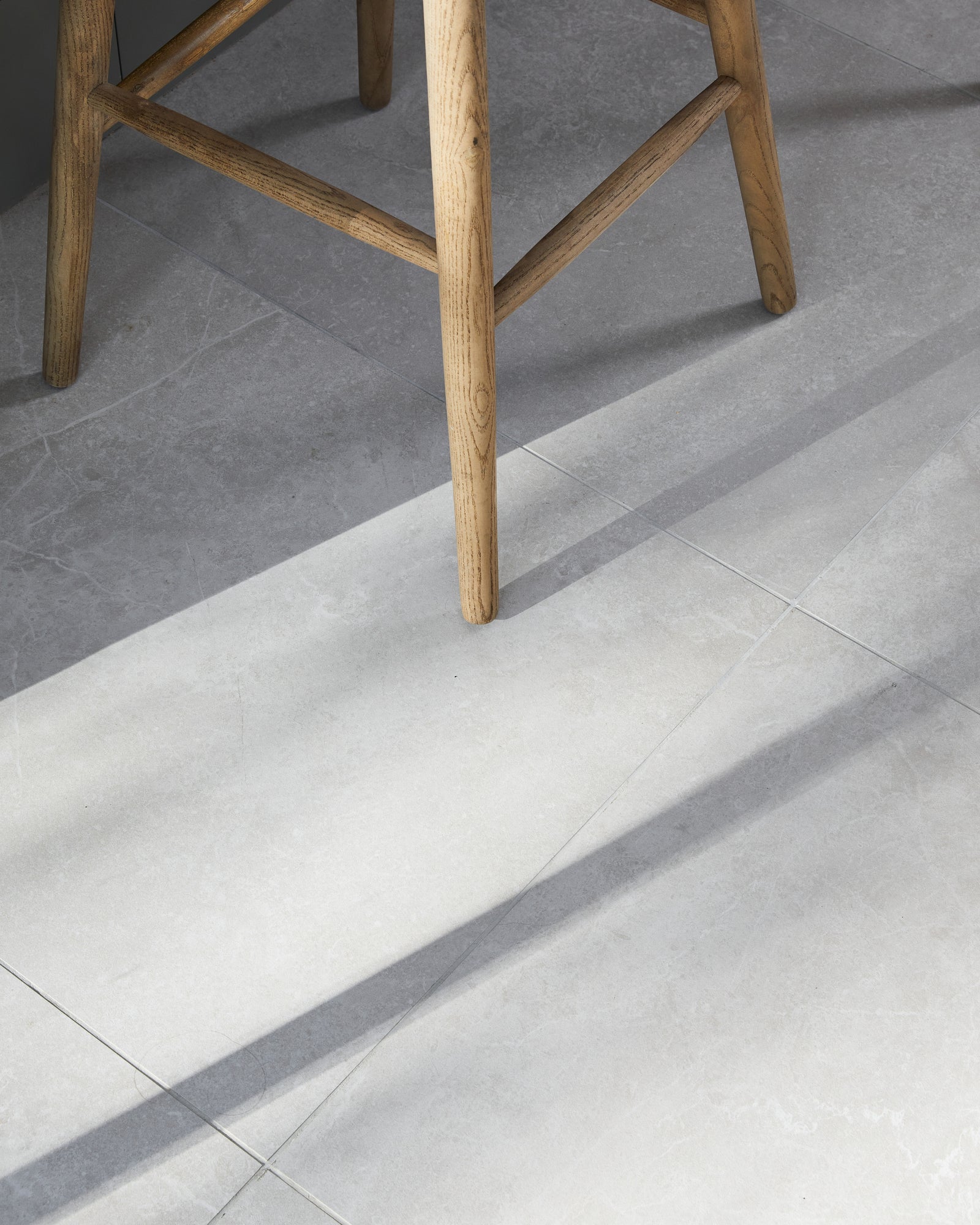 Close-up of Riverside Porcelain Perla floor tiles under an oak bar stool, raking sunlight picking out the stone texture and fine grout joints