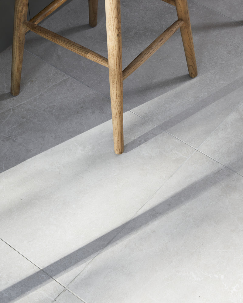 Close-up of Riverside Porcelain Perla floor tiles under an oak bar stool, raking sunlight picking out the stone texture and fine grout joints