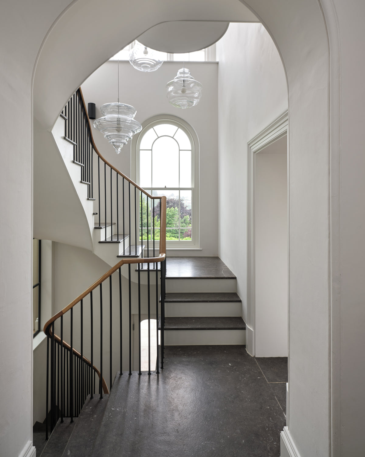 Royal Mink Limestone Tumbled Finish on the floor and stair treads of a double-height stair hall, seen through a plaster arch with glass pendants and an arched window