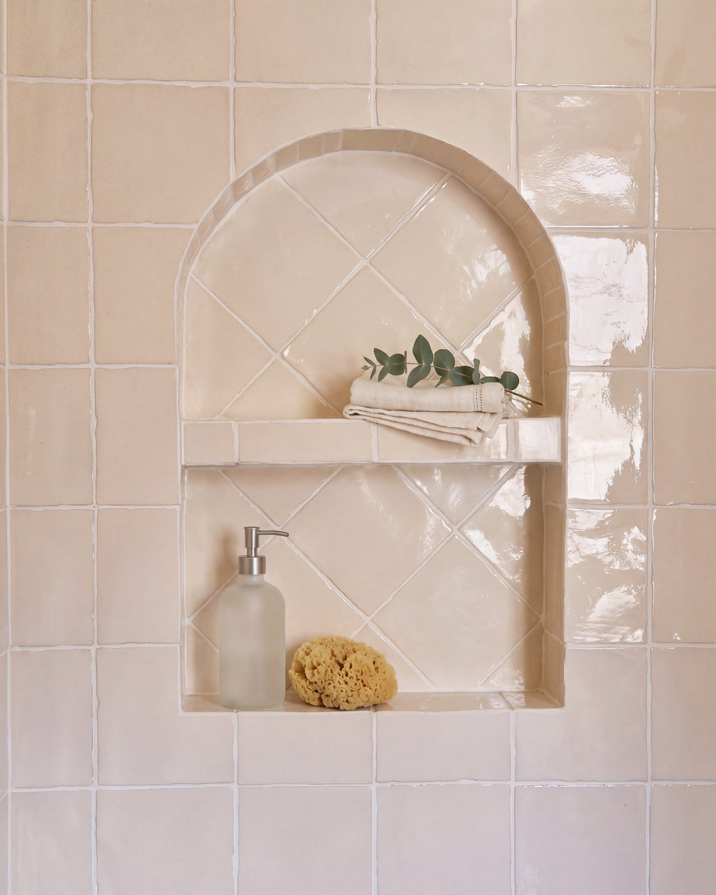 Rural Ceramic Buttermilk glossy square tiles across a bathroom wall around an arched recessed niche, the niche laid on the diagonal and styled with folded linen towels, eucalyptus, a frosted glass soap dispenser and a natural sponge
