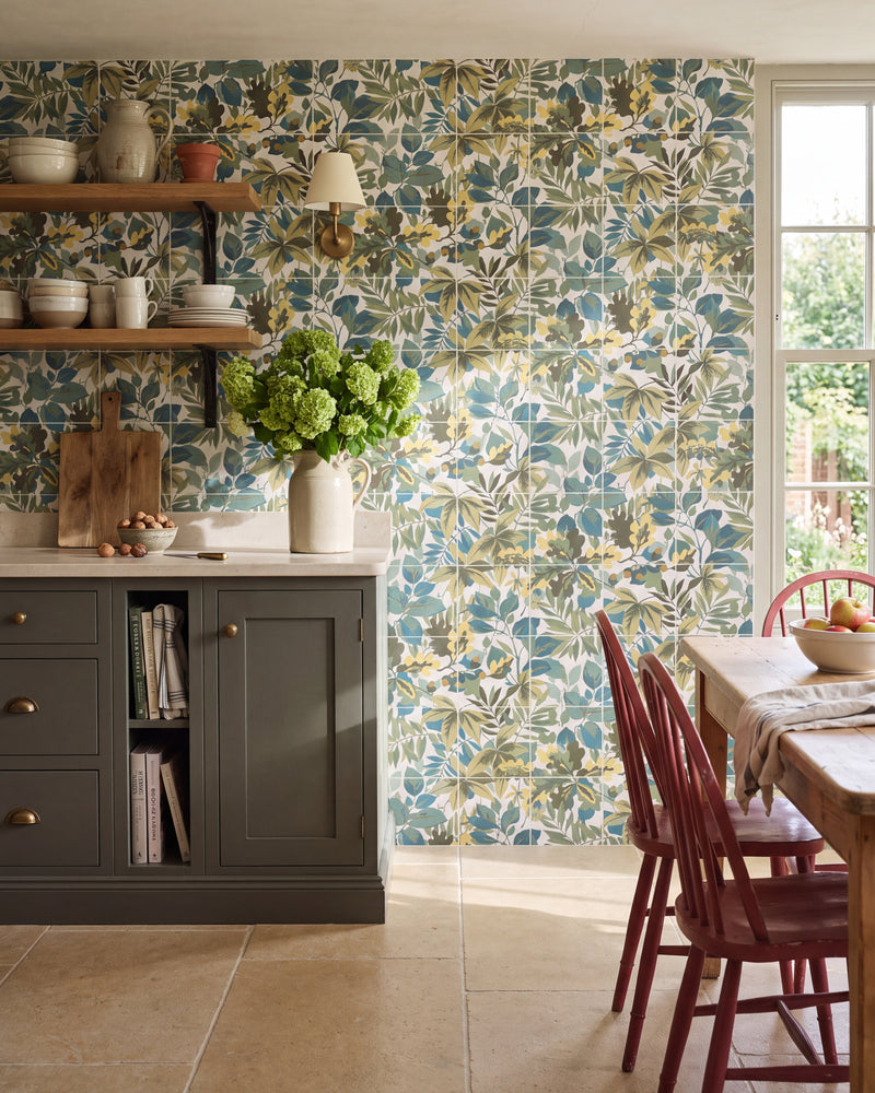 Sanderson Robin's Wood Sage Pattern tiled floor to ceiling as a kitchen splashback, with oak open shelving, a brass wall light, a stone worktop and red spindle-back chairs at a scrubbed pine table