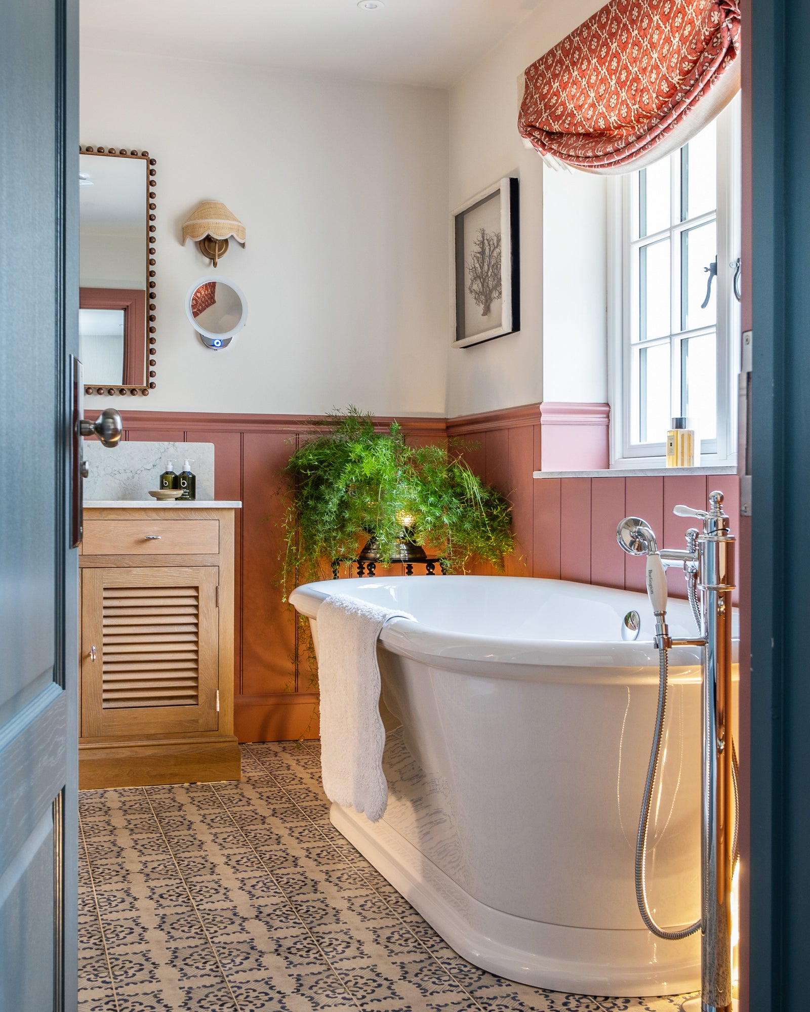 Sardinia Porcelain Tarricone running the length of a bathroom floor to a freestanding roll-top bath, with terracotta panelling, a pink window surround and a patterned Roman blind. Interior design by Fiona Doswell Interiors