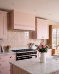 STUDIO Sanderson Anthos Pink Clay tiles as a kitchen splashback behind a black range cooker, set against blush pink shaker cabinetry, a marble worktop and oak dresser