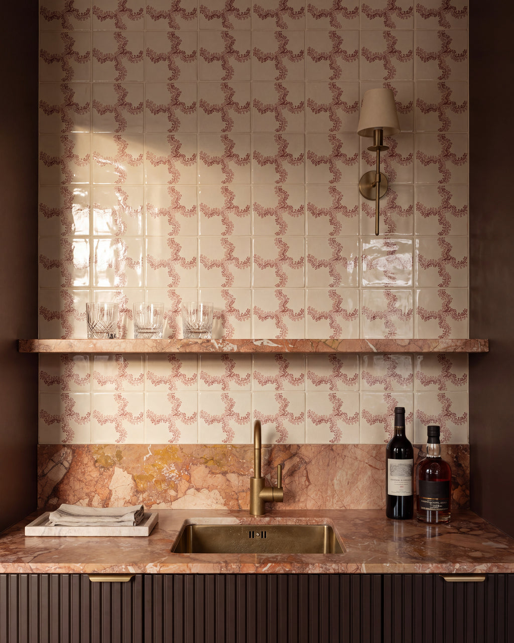 STUDIO Sanderson Oxbow Check Madder tiles behind a drinks bar, with a brass sink and tap, pink marble worktop and shelf, fluted cabinetry and a slim brass wall light