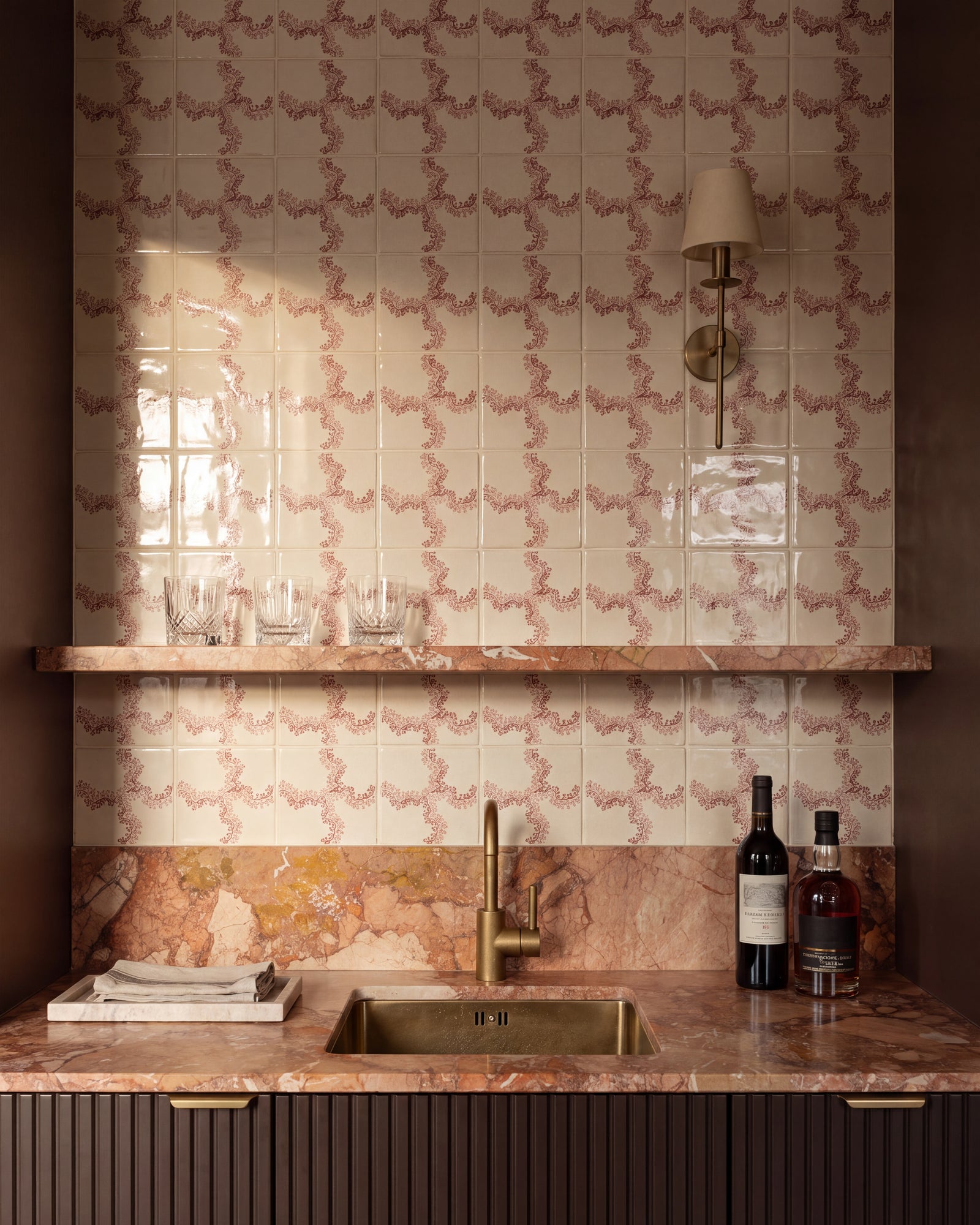 STUDIO Sanderson Oxbow Check Madder tiles behind a drinks bar, with a brass sink and tap, pink marble worktop and shelf, fluted cabinetry and a slim brass wall light