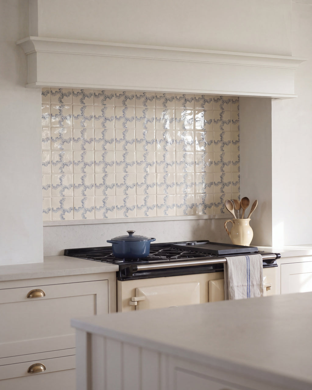 STUDIO Sanderson Oxbow Check Pottery Blue tiles as a kitchen splashback beneath a plastered chimney hood, above a cream range cooker with brass cup handles and a stone worktop