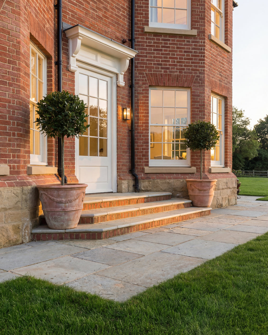 Trusloe Limestone Seasoned Finish paving and steps at the entrance to a red brick house, lit at dusk with bay trees in terracotta pots