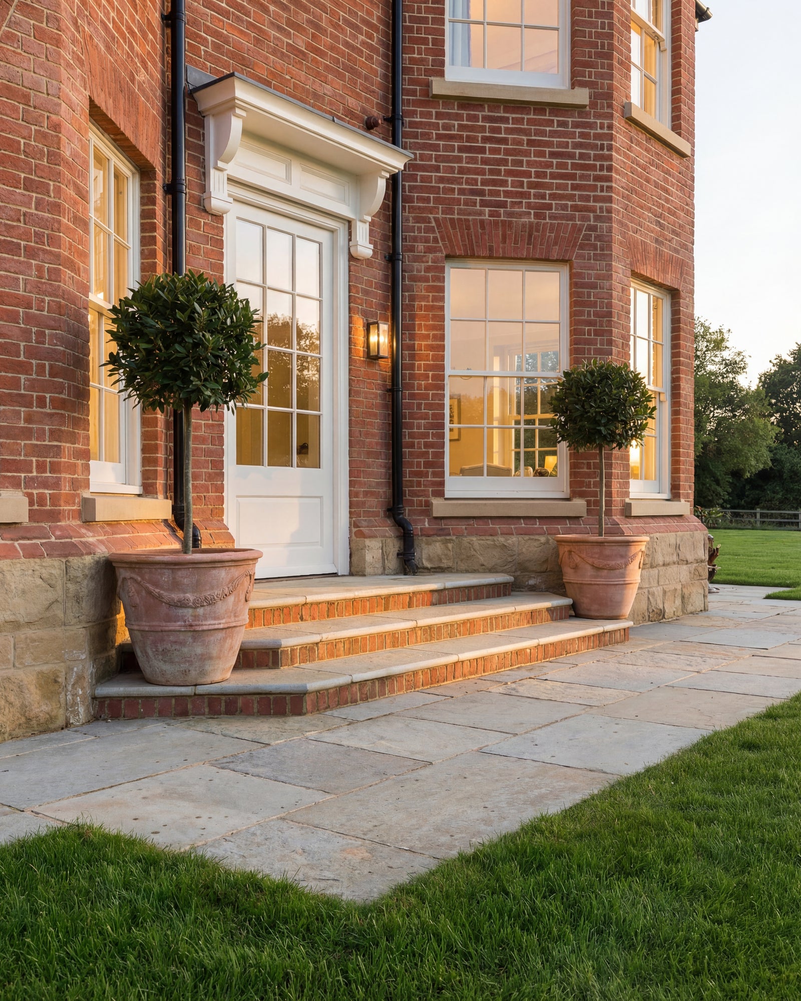 Trusloe Limestone Seasoned Finish paving and steps at the entrance to a red brick house, lit at dusk with bay trees in terracotta pots