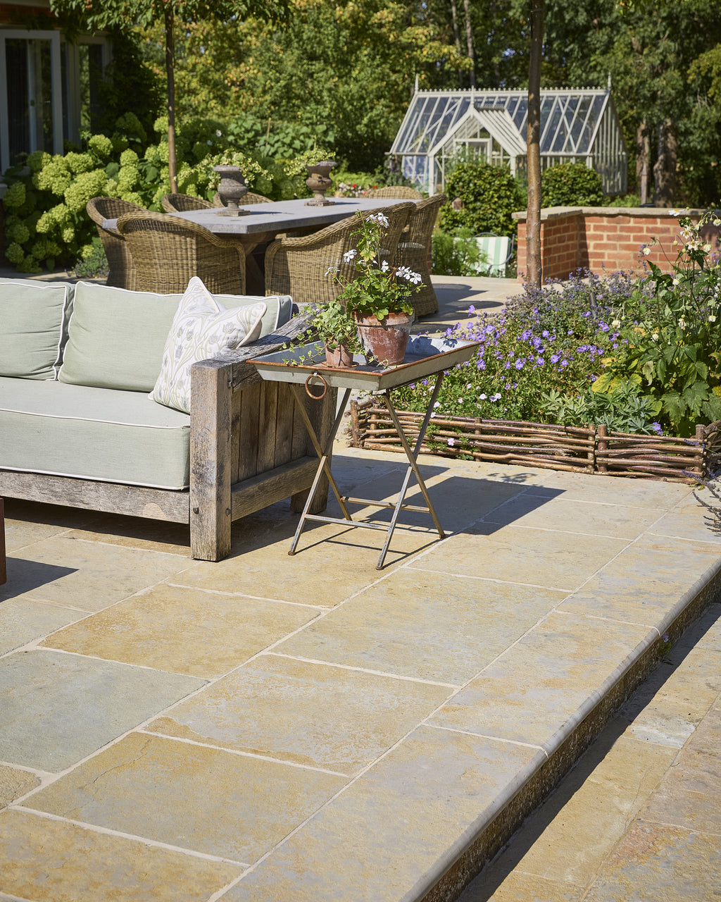 Trusloe Limestone Seasoned Finish paving on a sunlit garden patio with a weathered oak sofa, hydrangeas and a glasshouse beyond