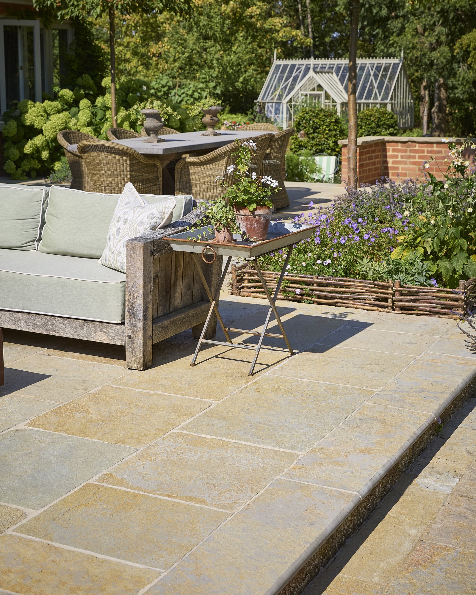 Trusloe Limestone Seasoned Finish paving on a sunlit garden patio with a weathered oak sofa, hydrangeas and a glasshouse beyond