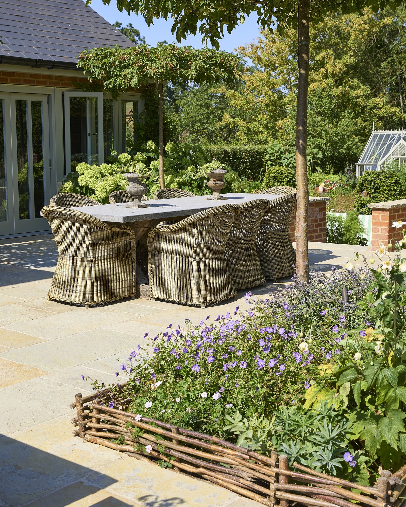Trusloe Limestone Seasoned Finish paving beneath a stone-topped dining table and wicker chairs, shaded by pleached crab apple trees
