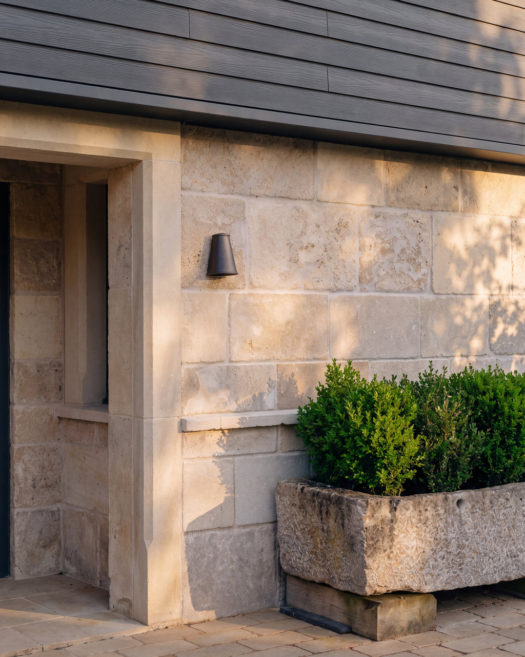 Vieux Bourgogne Limestone Ancient Finish as wall cladding around a covered entrance, lit low by an evening sun beneath grey timber boarding, with a boxwood-planted stone trough alongside