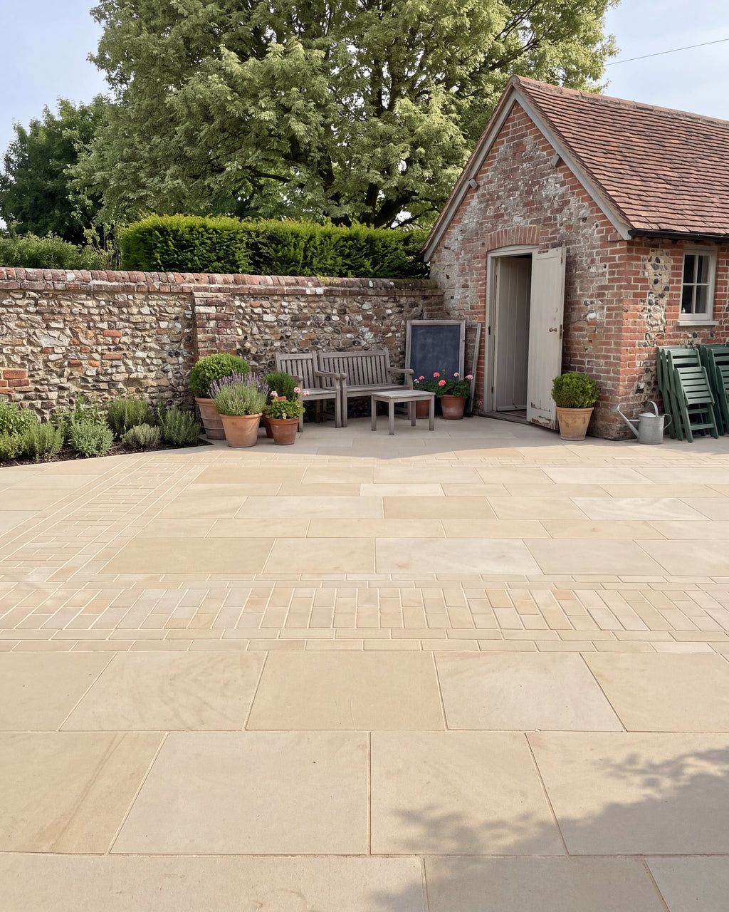 Wensley York Sandstone Honed paving across a walled courtyard beside a brick and flint outbuilding, with a weathered teak bench and terracotta pots of lavender