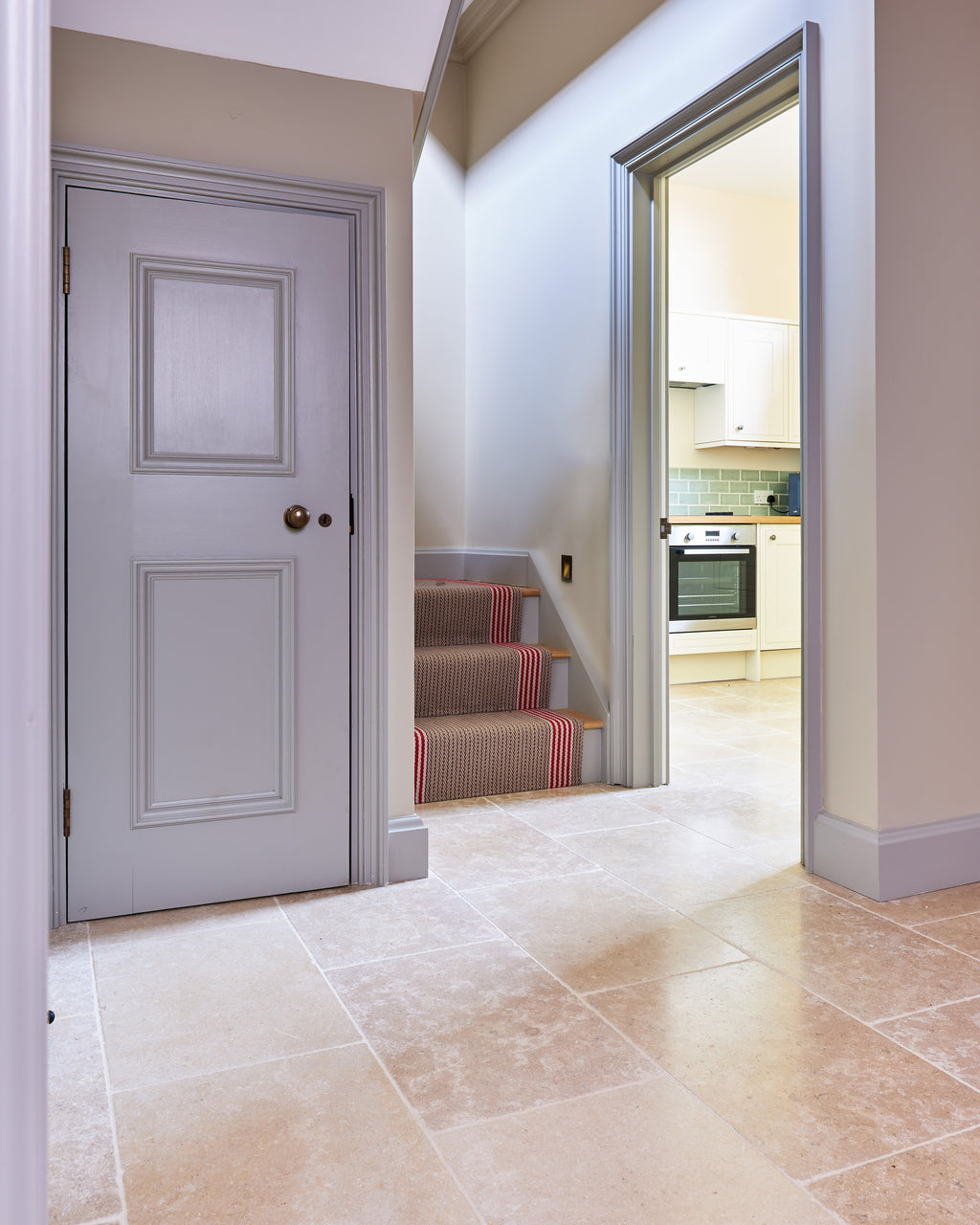 Wychwood Limestone Tumbled Finish flooring across a hallway with a panelled grey door, a red striped stair runner and an open doorway through to the kitchen